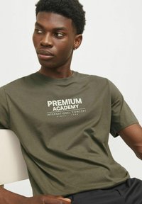 Olivno zelena bombažna majica s kratkimi rokavi z belim napisom "PREMIUM ACADEMY". Okrogla ovratnika, sproščen kroj, minimalna tekstura.