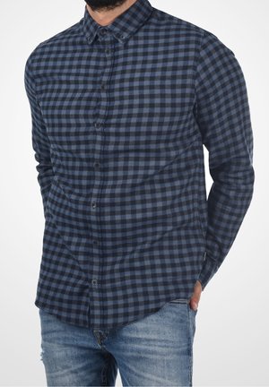 Mann trägt ein marineblaues und schwarzes kariertes Hemd mit Knöpfen, langen Ärmeln und hellblauen Jeans, eine Hand in der Tasche.