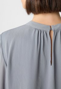 Hellgraue Bluse mit feinen vertikalen Falten, rundem Ausschnitt und einem Schlüssellochdetail auf der Rückseite. Weicher, strukturierter Stoff.