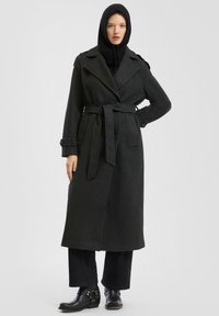 Zwarte wollen trenchcoat met een ceintuur in de taille, grote revers en knoopaccenten. gedragen over een zwarte gebreide capuchon en gecombineerd met zwarte broeken en laarzen.