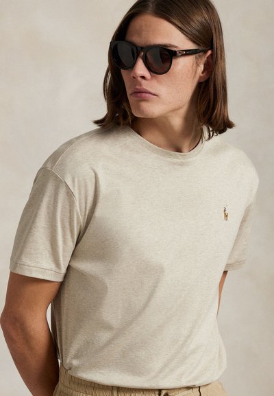 Lahek bež bombažni T-shirt s kratkimi rokavi in okroglim izrezom, z majhnim vezenim logotipom na prsih. Nošen z temnimi sončnimi očali.