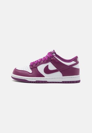 Baskets basses Nike blanches et violettes avec lacets, logo Swoosh violet et semelle en caoutchouc sur fond clair uni.