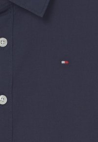 Chemise bleue marine à boutons avec des boutons blancs et un petit logo rectangulaire rouge et blanc sur la poitrine.