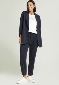 Eine lächelnde Frau in einem navyfarbenen Blazer und einer Hose, einer weißen Bluse und weißen Sneakers, die vor einem schlichten hellen Hintergrund steht.