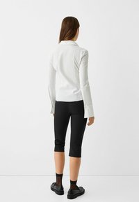 Woman standing facing away, wearing a white long-sleeve shirt, black cropped pants, sheer black socks, and black loafers.