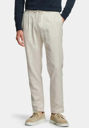 Homme portant un pantalon beige à cordon de serrage, des baskets décontractées beiges et une chemise noire à manches longues, debout avec une main dans la poche.