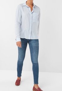 Camicia a righe blu chiaro con taschino sul petto, abbinata a jeans skinny blu scuro e scarpe slip-on rosse, su uno sfondo neutro.