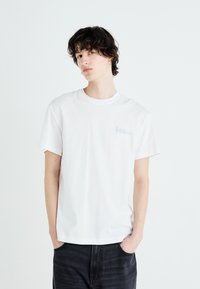 Weißes Baumwoll-T-Shirt mit rundem Halsausschnitt und kurzen Ärmeln. Dezentes blaues Logo auf der linken Brust. Kombiniert mit schwarzen Jeanshosen.