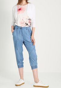 Cárdigan blanco ligero sobre una blusa de estampado floral, combinado con pantalones azules relajados que tienen una cintura con cordón y puños elásticos. Zapatillas blancas.