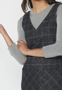 Dark plaid dress layered over a grey long-sleeve shirt. The dress features a V-neck and textured fabric with a structured fit.