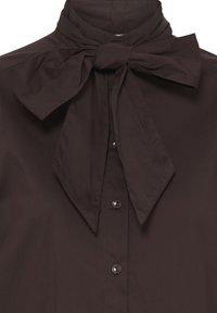 Chemise boutonnée marron foncé avec un grand nœud noué au col, présentée de la poitrine vers le haut sur un fond uni.