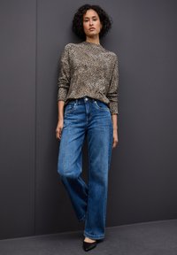 Pull en imprimé léopard beige et marron, associé à un jean large bleu clair et des escarpins noirs à bout pointu sur un fond gris foncé.