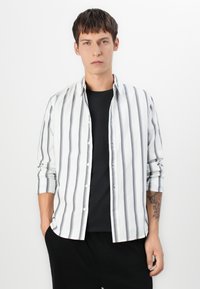 Joven con cabello corto castaño que lleva una camisa blanca de botones con rayas verticales negras sobre una camiseta negra, con las manos en los bolsillos.