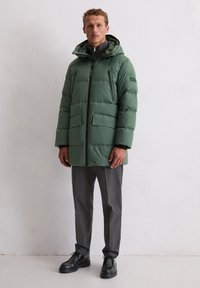 Kurtka puchowa w stonowanym zielonym kolorze, z kapturem, zamkiem błyskawicznym, dwoma przednimi kieszeniami i gładką teksturą. Połączona z szarymi spodniami i czarnymi butami.