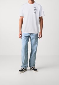 Personne portant un t-shirt blanc à manches courtes avec un graphique noir sur la poitrine, un jean léger bleu clair ample, et des baskets noires et blanches sur fond uni.