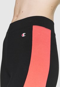 Närbild av svarta leggings med ett brett korallfärgat rand på sidan och en liten Champion-logotyp nära midjan.