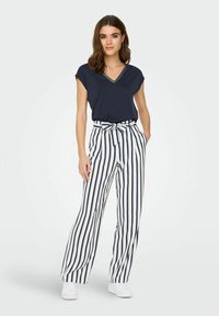 Haut bleu marine à manches courtes avec un col en V et des détails argentés, associé à un pantalon taille haute à rayures verticales blanches et bleu marine, et des baskets blanches.