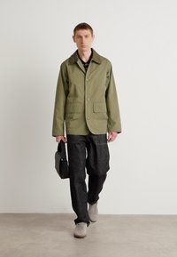 Olive green, collared jacket with two front pockets, paired with dark trousers and beige sneakers. The model carries a black bag.