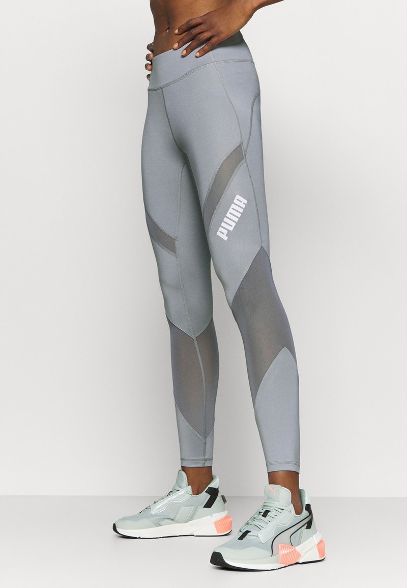 Leggings de sport gris avec des inserts en maille, taille haute et logo Puma blanc sur le côté, associés à des baskets bleu clair et noires.