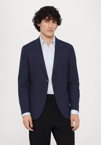Donkerblauwe stropdasblazer van gestructureerde stof, met een sluiting met één knop en twee voorzakken. Gecombineerd met een lichtblauw shirt.