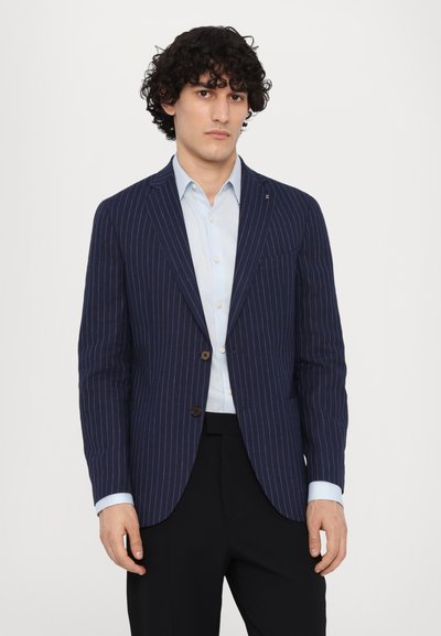 Blazer rayé marine en tissu texturé, avec une fermeture à un bouton et deux poches avant. Associé à une chemise bleu clair.