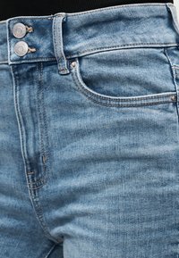 Ανοιχτό μπλε τζιν με διπλά μεταλλικά κουμπιά και εμφανή ραφή στην μπροστινή τσέπη, φορεμένα με μαύρο τοπ.