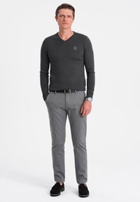Maglione grigio a maniche lunghe con scollo a V e un piccolo logo, abbinato a pantaloni chinos grigi chiari e scarpe slip-on nere, in piedi su uno sfondo bianco.