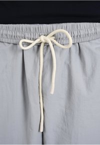 Pantalon gris clair avec une ceinture élastique et un nœud de cordon blanc noué sur un fond noir.