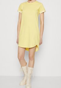 Robe jaune à manches courtes avec un col rond et des fentes sur les côtés. Texture lisse et coupe décontractée, associée à des bottes à lacets beiges.