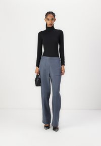 Schwarzes Rollkragenoberteil; graue weite Hose mit Fronttaschen; Modell hält eine kleine schwarze Tasche; glänzende schwarze High-Heels.