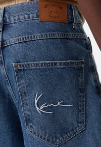 Μπλε τζην παντελόνι με δερμάτινη ετικέτα που φέρει ανάγλυφο το όνομα της μάρκας. Λευκή κεντητή λεπτομέρεια υπογραφής στην πίσω τσέπη. Κλασικός σχεδιασμός.