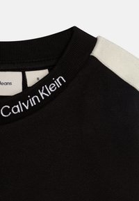 Crni pulover s rebrastim ovratnikom, na kojem se nalazi bijela Calvin Klein vezna oznaka, s kremastim ramenim krpicama.