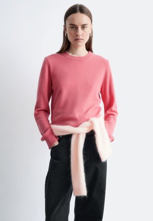 Rosa uldtrøje med rund hals, kombineret med mørke jeans. Et blødt, lyserødt tørklæde er bundet omkring taljen. Glat tekstur, afslappet design.