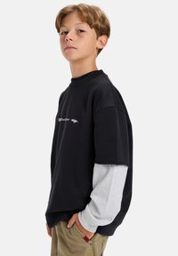 Ragazzino con capelli castano chiaro, vestito con maglie lunghe sovrapposte nere e grigie e pantaloni khaki, in piedi con una mano in tasca su uno sfondo bianco.
