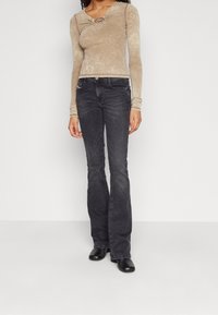 Beżowy top z długim rękawem o skręconym przodzie i okrągłym akcentem, w zestawieniu z ciemnymi dżinsami w stylu dzwonów oraz czarnymi botkami na obcasie.