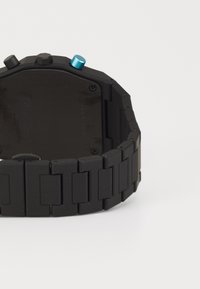 Montre noire avec finition mate, dotée d'un boîtier rectangulaire, de maillons audacieux, et de deux boutons ; un bleu, un gris en haut.