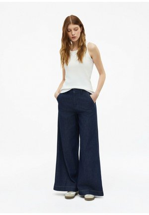 Frau mit langen Haaren, die ein weißes ärmelloses Oberteil, dunkle Jeans mit weitem Bein und weiße Sneakers trägt, steht mit den Händen in den Taschen.