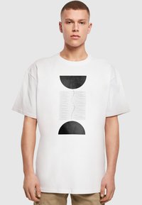 Hvid bomulds t-shirt med et sort grafisk design bestående af to halvcirkler og bølgede linjer imellem, korte ærmer og en afslappet pasform.