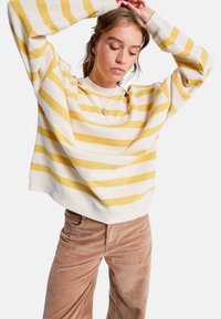 Cremfarbener und gelb gestreifter Sweatshirt mit langen Ärmeln und Rundhalsausschnitt. Der Stoff wirkt weich, mit lässigem Schnitt und sichtbarem Logo. Braune Cordhosen.