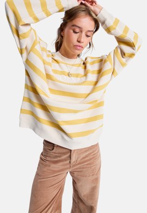 Crème en gele gestreepte sweatshirt met lange mouwen en ronde hals. De stof lijkt zacht, met een relaxte pasvorm en zichtbaar logo. Bruine korduroy broek.