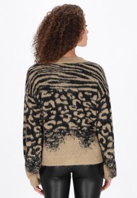 Beige, oversize pulóver fekete állatmintával, kötött textúrával és flitteres díszítéssel a ujján. A hátsó nézeten ejtett vállak láthatók.