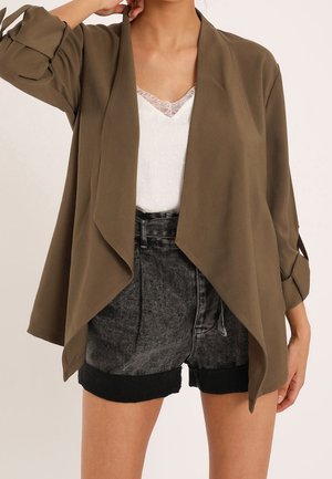 Kobieta ubrana w oliwkowo-zieloną marynarkę bez zapięcia na biały top z koronkowym wykończeniem i czarne, wysokiego stan, podwinięte szorty dżinsowe, z ręką podniesioną do głowy.