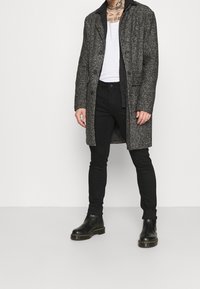 Abrigo de pata de gallo en blanco y negro con cuello de muesca, llevado sobre una camisa sin mangas blanca, combinado con pantalones negros slim-fit y botines negros.