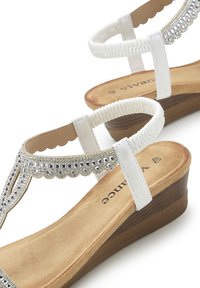 Weiße Keilsandaletten mit einem strukturierten Riemen und silbernen Akzenten, die mit Strasssteinen verziert sind, und einer hellbraunen Innensohle auf einer braunholzähnlichen Sohle.