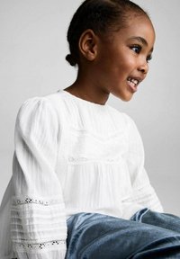 Witte blouse met textuurstof, kantaccenten en pofmouwen. Het kind draagt een blauwe fluwelen outfit en zit tegen een neutrale achtergrond.