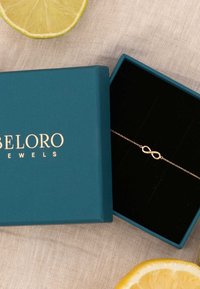 Bracelet en or en forme d'infini à l'intérieur d'une boîte turquoise avec du texte doré. Intérieur en velours noir doux. Entouré de tranches de citron sur une surface texturée.