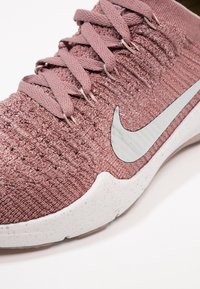 Nike athletic shoe in light pink knit material with textured surface, featuring white sole and silver swoosh logo, round toe, and lace-up design.