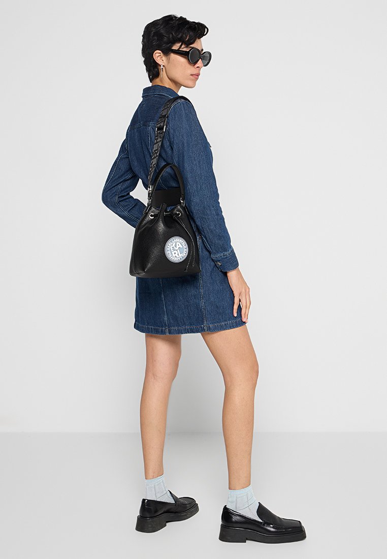Schwarze Leder-Eimer-Tasche mit einem runden Logo-Patch, strukturiertem Riemen und gerafftem oberen Teil. Modell trägt ein Denim-Kleid und schwarze Schuhe.