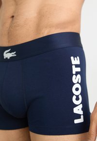 Navyblauwe boxershorts van zacht materiaal, met een wit Lacoste-logo op de zijkant en een comfortabele elastische tailleband.