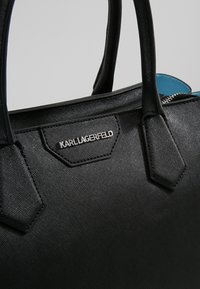 Czarna torebka o teksturowanej powierzchni z gładkimi uchwytami, srebrnym logo oraz akcentem niebieskiego koloru na suwakach. Uformowana w zorganizowany kształt z kanciastymi detalami.